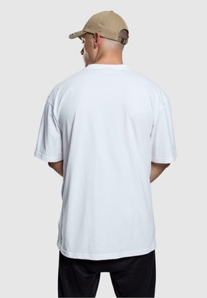 Camiseta de algodón blanca de manga corta, corte relajado, diseño liso, vista desde la parte posterior. Combinada con una gorra de béisbol beige.