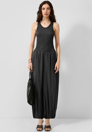 Woman wearing a sleeveless black top and loose black pants holding a black mesh bag, standing against a plain light gray background.