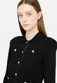 Giovane donna con capelli castani lisci, che indossa una giacca nera con bottoni dorati e orecchini dorati a forma di cuore, su uno sfondo bianco.