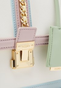 Gros plan sur un sac en cuir pastel avec une chaîne en or et un fermoir en or gravé de "ALDO" sur un matériau blanc texturé.