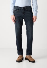 Σκούρα μπλε τζιν με ίσια γραμμή, πεντατσέπη σχέδιο, διακριτές ραφές, φορεμένα με ανοιχτό καφέ μοκασίνια.