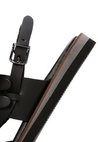 Sandalia de cuero negro con correa ajustable y hebilla plateada; la suela plana presenta acentos en capas de color negro, gris y marrón.