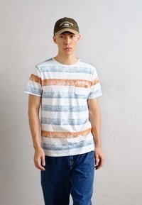 Camiseta de algodón a rayas con líneas horizontales azules y naranjas y un bolsillo en el pecho izquierdo. Se lleva con jeans de mezclilla azules y una gorra verde.