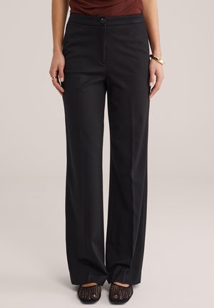 Frau trägt schwarze, weit geschnittene Hosen, braunes Oberteil, schwarze Schuhe mit offenem Maschenmuster, goldenes Armband und Ring, steht vor einem einfarbigen Hintergrund.