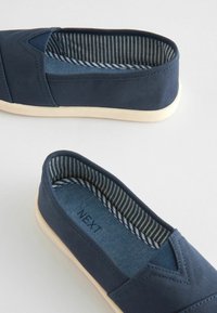 Baskets slip-on en toile marine avec une semelle légère ; l'intérieur présente une doublure rayée bleue et blanche. Comprend un discret design en forme de V à l'avant.