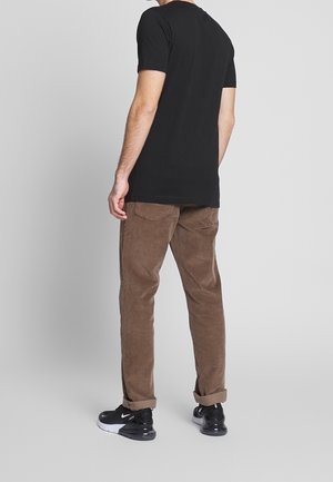 Mann steht mit dem Rücken zur Kamera, trägt ein schwarzes T-Shirt, braune Cordhosen mit hochgekrempelten Säumen und schwarz-weiße Nike-Sneaker.