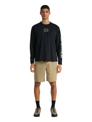 Man draagt een zwart shirt met lange mouwen met witte tekst op borst en mouw, beige cargoshort, zwarte sokken en beige sneakers en staat met het gezicht naar voren.