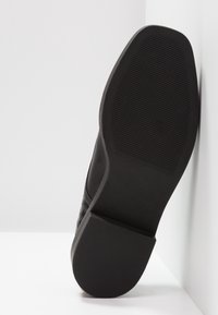 Semelle de sneaker en caoutchouc noir avec des motifs de grip texturés, présentant un talon légèrement surélevé et une surface latérale lisse.