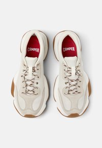 Un par de zapatillas chunky blancas y beige con suelas marrones, cordones estampados y plantillas rojas con la marca "CAMPER", vistas desde arriba.
