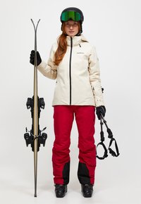 Kvinde i beige skijakke og røde bukser, iført hjelm og skibriller, holder skis og skistave mod hvid baggrund.