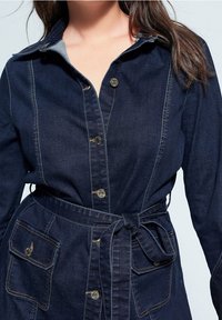 Robe en denim bleu marine avec un col, des boutons sur le devant, deux poches plaquées, une taille ceinturée et des détails de couture contrastés.