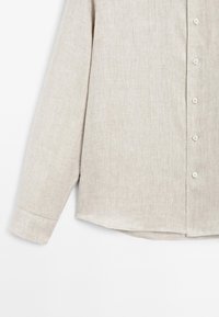 Beige long-sleeve button-up shirt with a textured fabric. Features a classic collar, rounded hem, and subtle stitching along cuffs and buttons.