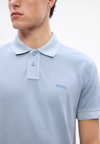 Svetlomodrá polokošeľa z texturovanej tkaniny s klasickým golierom, dvoma gombíkmi a malým logom "BOSS" v zodpovedajúcej modrej farbe.