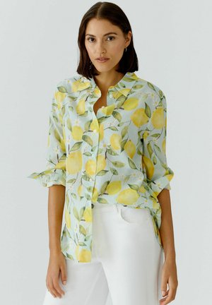 Vrouw met donker haar tot op schouderlengte, die een lichtblauw overhemd met gele citroenprint en een witte broek draagt tegen een effen achtergrond.