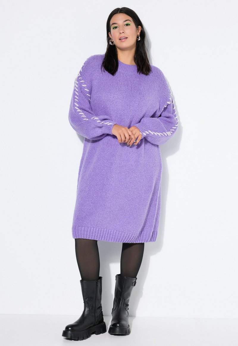 Abito a maglia viola con maniche lunghe, polsini e bordi a coste, caratterizzato da dettagli di cucitura bianca lungo le maniche. Indossato con stivali neri.