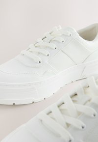 Baltos sportinės bateliai, pagaminti iš lygios odos, apvalia nosimi, su šešiais skylučių raišteliais, stambiu baltu padu ir subtiliu toniniu siuvimu.