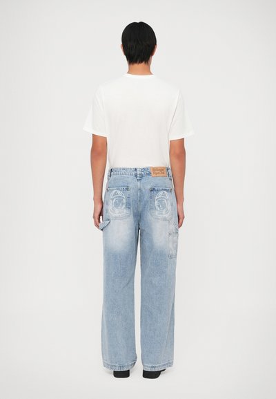 Jeans ancho de color azul claro con un lavado desgastado, sutil estampado de astronauta en los bolsillos y un parche de cuero marrón en la cinturilla.