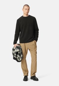 Czarny sweter z dzianiny, beżowe spodnie i czarne buty. Trzyma wzorzysty szary i czarny szal. Prosty design z luźnym krojem.