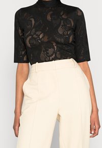 Schwarzes Spitzentop mit kurzen Ärmeln und hohem Kragen, verziert mit floralen Mustern und transparenten Elementen, kombiniert mit hochtaillierten, cremefarbenen Hosen.