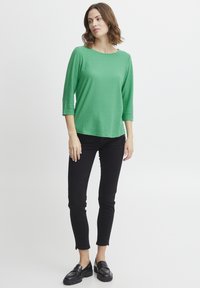 Zelený textúrovaný top s okrúhlym výstrihom a trojštvrťovými rukávmi, v kombinácii s čiernymi slim-fit nohavicami a čiernymi mokasínami.