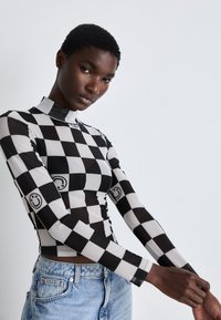 Long-sleeve, fitted top with a black and white checkerboard pattern. Features a high collar and smiley face accents on the sleeves.