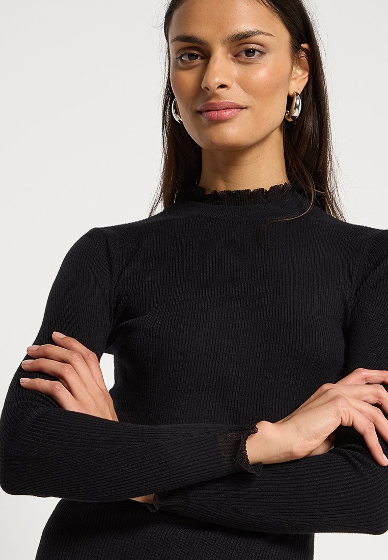 Czarny, prążkowany top z długim rękawem o wysokim kołnierzu, z falbaną na krawędzi i dopasowaną sylwetką. Materiał jest miękki i elastyczny.
