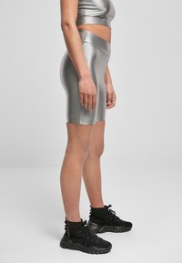 Kobieta w srebrnych, wysokich szortach rowerowych i dopasowanej crop topie, w połączeniu z czarnymi sportowymi butami na sznurówki na jednolitym tle.