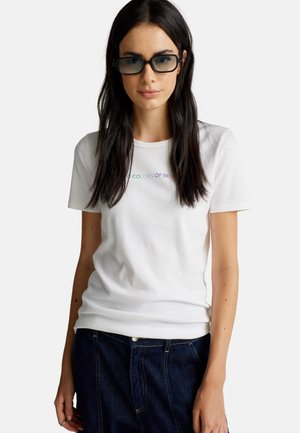 Donna con lunghi capelli scuri che indossa occhiali da sole rettangolari scuri, una maglietta bianca con testo multicolore sbiadito e jeans blu scuri.