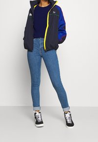 Marinblå och livfull blå jacka med gul dragkedja, kombinerad med blåa skinny jeans och svarta högklackade sneakers med vita snören.