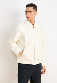 Beige zip-up jacket with a collar, side pockets, and a textured fabric. Paired with black cargo pants, presented against a plain background.