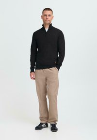 Sweat à zip noir avec un col haut, associé à un pantalon beige. Le modèle se tient debout dans des baskets noires, montrant une coupe décontractée.