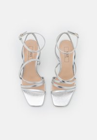 Srebrne metaliczne sandały na wysokim obcasie z wieloma cienkimi paskami, kwadratowym noskiem i gładką fakturą. Podeszwa w kontrastującym cielistym kolorze.