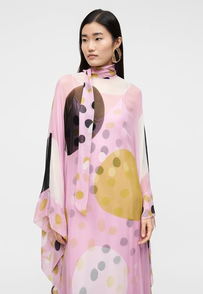 Žena s dlhými tmavými vlasmi má na sebe priesvitné ružové šaty s veľkými farebnými bodkami a veľké zlaté kruhové náušnice na jednoduchom pozadí.