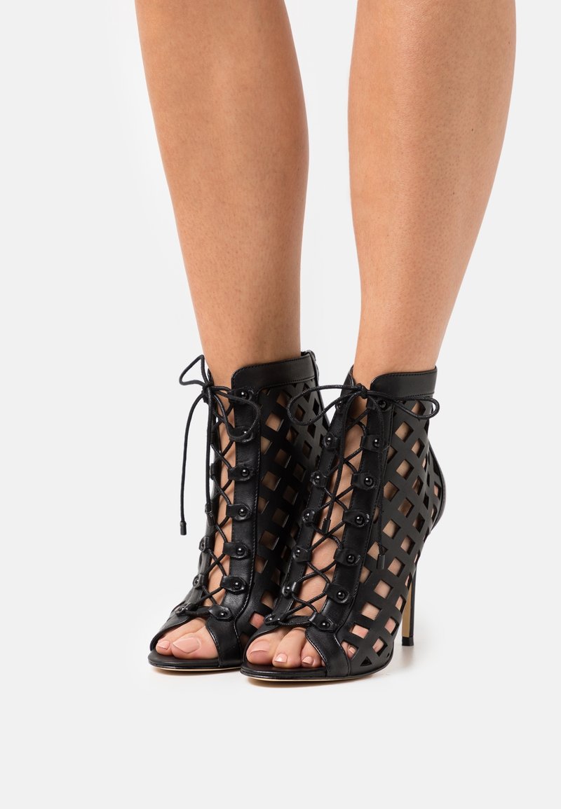 Bottines noires à talons hauts, ouvertes à l'avant, avec un design ajouré en treillis et une fermeture à lacets à l'avant, portées aux pieds d'une personne sur un fond blanc.