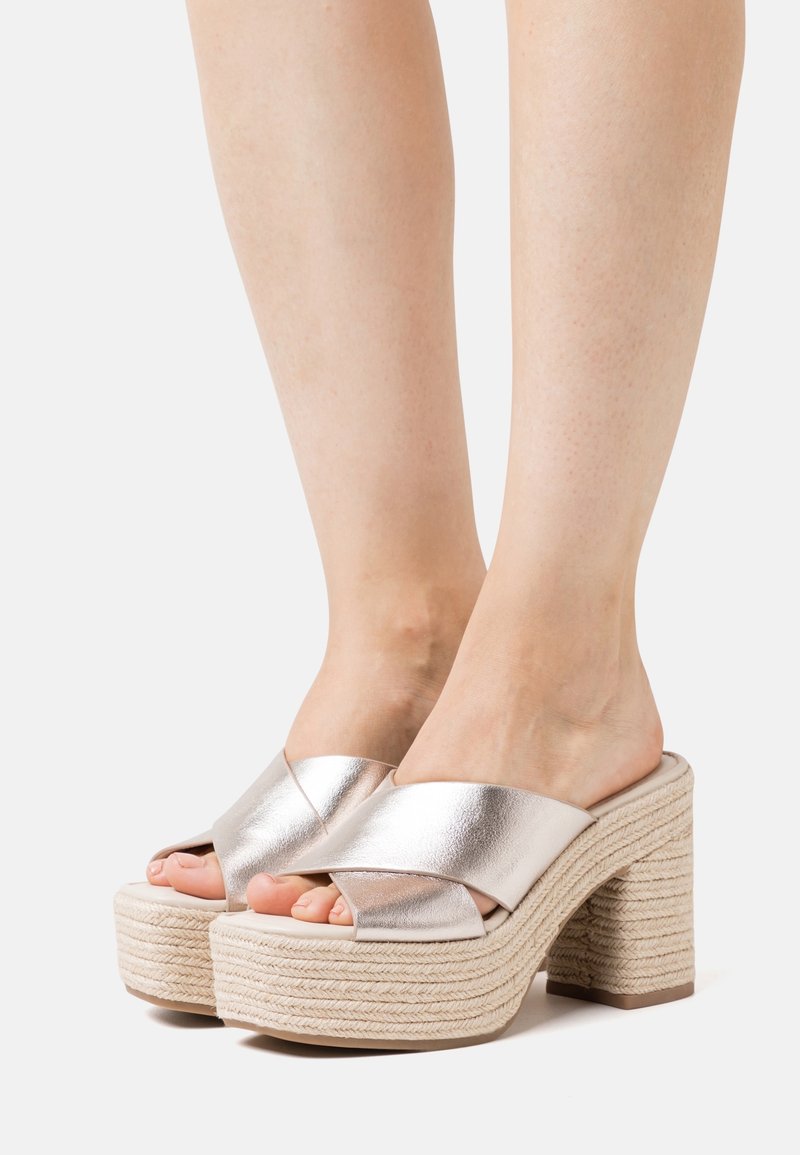 Sandalias de plataforma de metal plateado con tiras cruzadas, suela texturizada estilo yute y tacón cuadrado. Parte trasera cerrada con diseño de punta abierta.