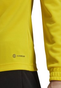 Maglietta sportiva gialla a maniche lunghe con una superficie testurizzata e un logo nero "AEROREADY" vicino all'orlo inferiore. Le maniche presentano polsini arricciati.