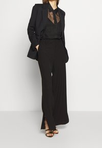 Zwarte op maat gemaakte blazer met textuur op de revers, doorschijnend zwart blouse en wijde zwarte trousers. Strappy zwarte sandalen complementeren de outfit.