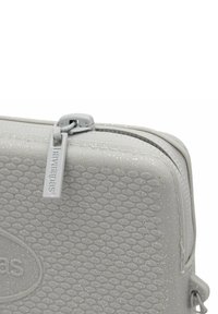 Funda texturizada gris con cierre de cremallera, que presenta una superficie con un patrón y un sutil brillo. Incluye una etiqueta con el logo "Havaianas".