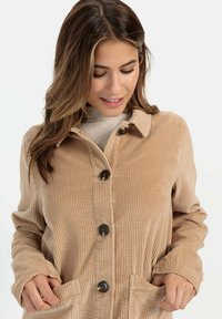 Beige Kordjacke mit Kragen, Knopfverschluss und seitlichen Taschen. Sie verfügt über eine vertikale gerippte Textur und runde, dunkle Knöpfe.