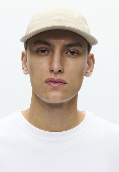 Νέος άνδρας με ουδέτερη έκφραση που φοράει μπεζ καπέλο με κέντημα "W.W." και λευκό πουλόβερ με στρογγυλή λαιμόκοψη, πάνω σε απλό ανοιχτόχρωμο φόντο.
