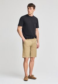 T-shirt nera a maniche corte con tasca, abbinata a pantaloni corti color khaki. I pantaloni corti hanno un taglio dritto e sono indossati con una cintura nera e scarpe marroni.