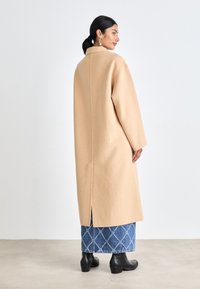 Cappotto lungo beige con una vestibilità rilassata, realizzato in misto lana. Presenta una finta a dietro per il movimento. Indossato sopra una gonna in denim trapuntato e stivali neri.