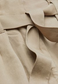 Beige látka se strukturovaným tkaním, s látkovými stuhami, které jsou volně zavěšeny v působivém uspořádání. Jemný lesk dodává hloubku materiálu.