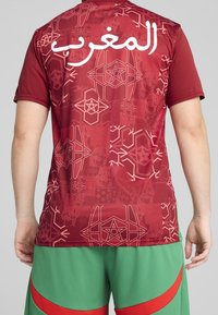 قميص رياضي باللون البني المحمر يتزين بأنماط هندسية وكلمة "المغرب" باللون الأبيض. القماش ناعم مع أكمام قصيرة.