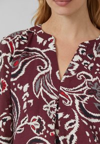 Burgunderfarbene Bluse mit V-Ausschnitt und aufwendigen weißen, grauen und roten Blumenmustern. Weicher Stoff mit leichtem Textur und Raffung an den Schultern.