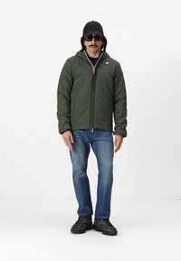 Grøn polstret jakke med lynlås foran, hætte og elastiske manchetter, parret med blå jeans og sorte sneakers. Modellen står foran en hvid baggrund.