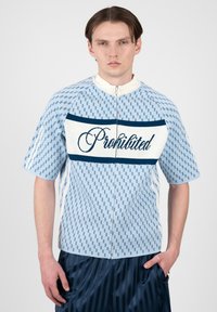 Jeune homme portant une chemise zippée bleu clair à motifs avec le texte « Interdit » sur la poitrine et un pantalon rayé bleu foncé.
