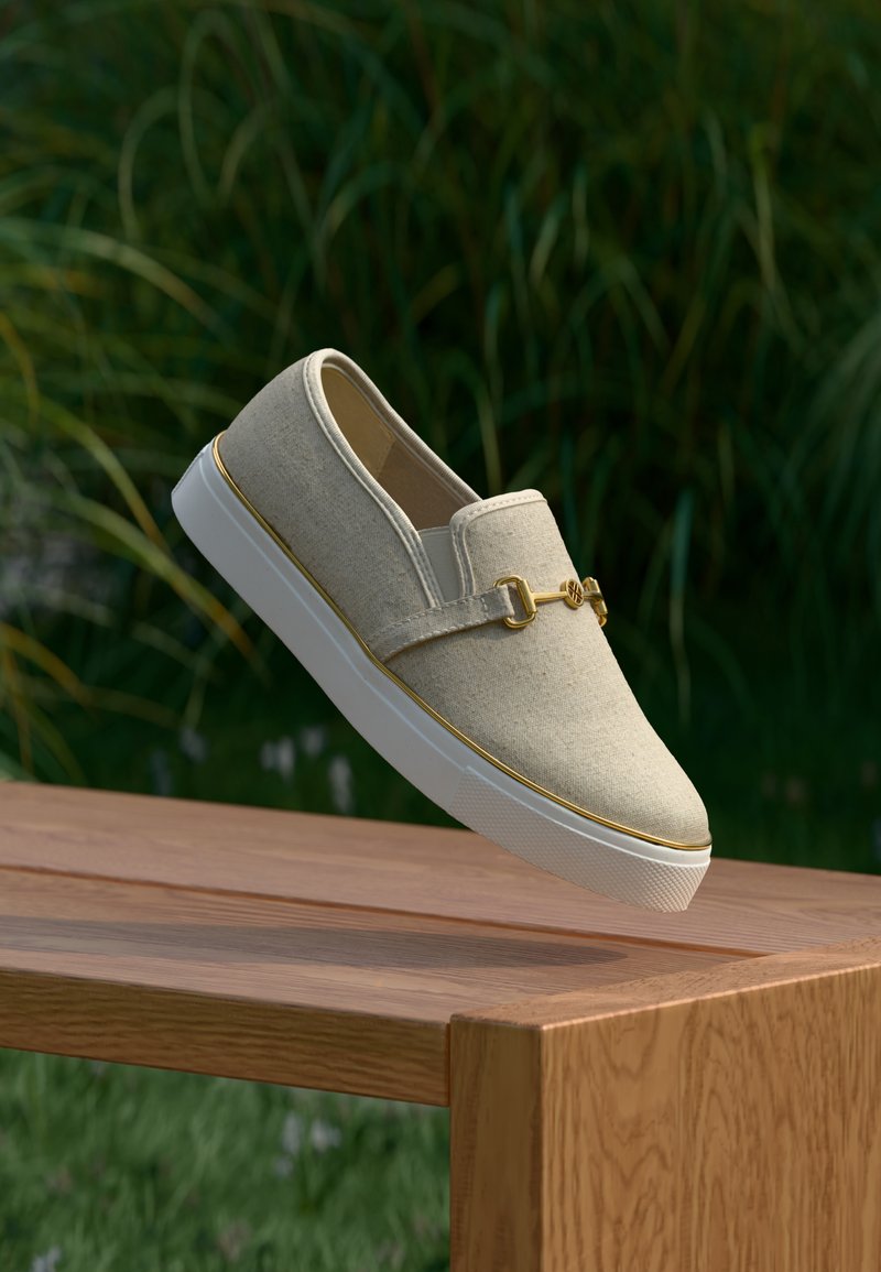 Chaussure beige à enfiler avec semelle blanche et détail boucle dorée, positionnée au-dessus d'un banc en bois en plein air avec des plantes vertes en arrière-plan.