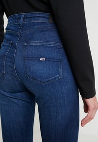 Acercamiento de una persona que lleva puestos unos pantalones vaqueros ajustados de color azul oscuro y una camiseta de manga larga negra, mostrando el bolsillo trasero con un pequeño detalle de logo.
