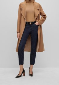 Lång kamelrock över en beige polotröja, mörkblå jeans, svarta högklackade skor, med ett svart bälte med ett guldaccenter.